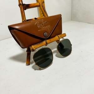 Op de afbeelding: Ray-Ban zonnebril met een bruine leren etui, gepresenteerd op een bamboe standaard. De etui heeft het Ray-Ban logo en de tekst "SUN GLASSES MADE IN U.S.A.". De zonnebril heeft donkere glazen en een gouden montuur.