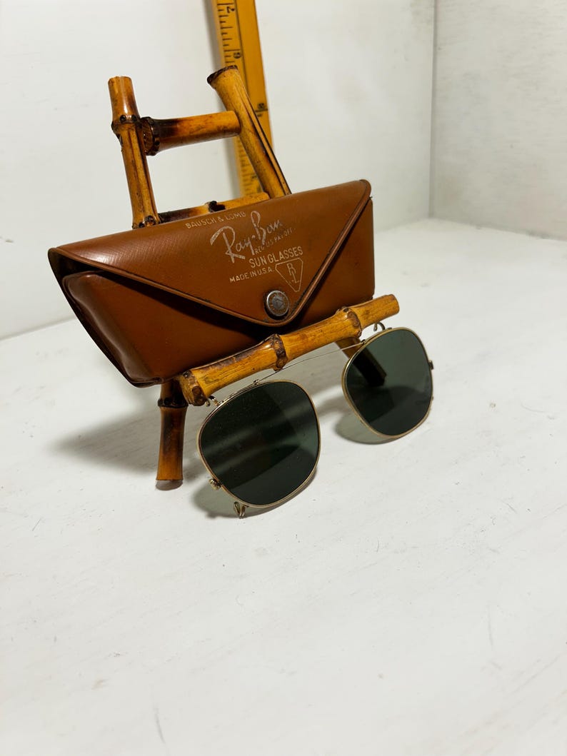 Op de afbeelding: Ray-Ban zonnebril met een bruine leren etui, gepresenteerd op een bamboe standaard. De etui heeft het Ray-Ban logo en de tekst "SUN GLASSES MADE IN U.S.A.". De zonnebril heeft donkere glazen en een gouden montuur.