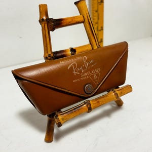 Op de afbeelding: Een bruine leren Ray-Ban zonnebril etui met de merknaam en de tekst "MADE IN U.S.A." weergegeven. De etui rust op een bamboe standaard. De standaard is gemaakt van aan elkaar gemaakte bamboestokken.