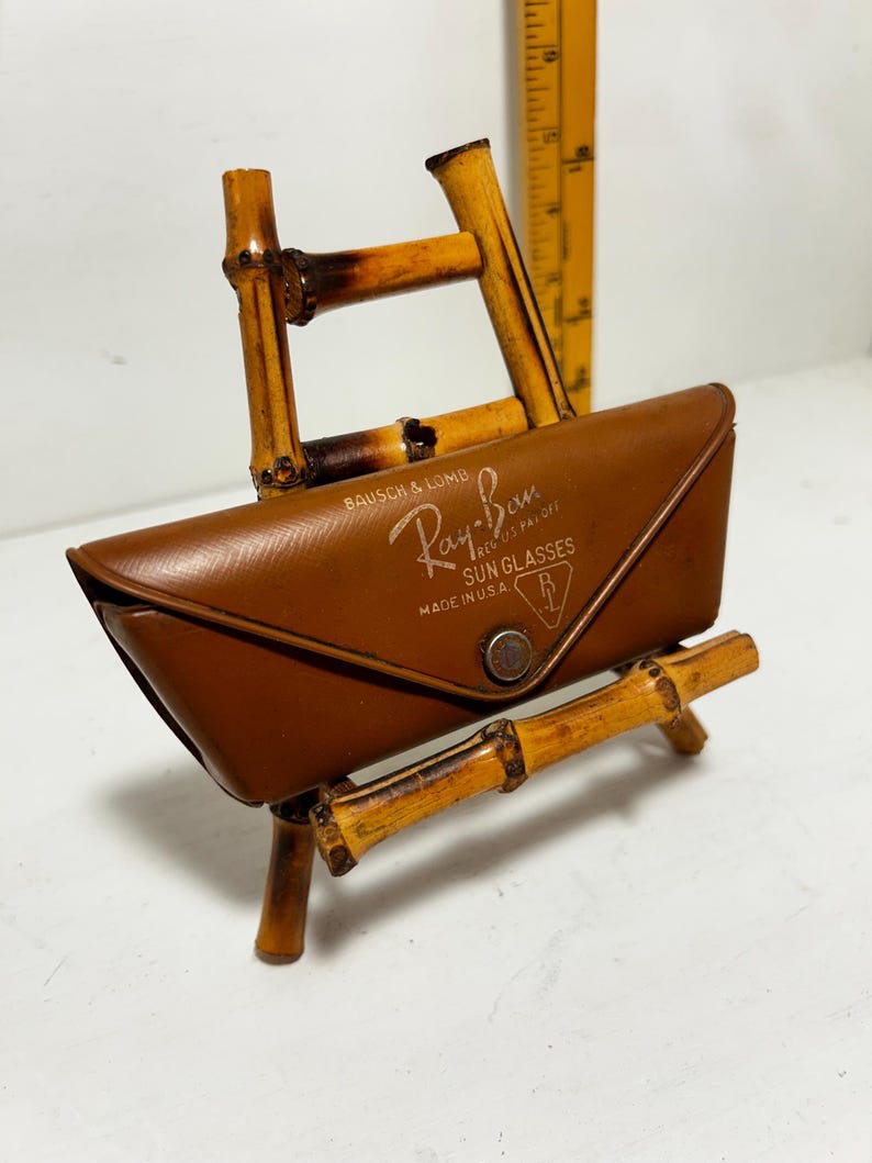 Op de afbeelding: Een bruine leren Ray-Ban zonnebril etui met de merknaam en de tekst "MADE IN U.S.A." weergegeven. De etui rust op een bamboe standaard. De standaard is gemaakt van aan elkaar gemaakte bamboestokken.