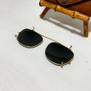 Op de afbeelding: Vintage clip-on zonnebril met donkere glazen en een goudkleurig montuur. De zonnebril ligt op een wit oppervlak, met een bruine leren etui op de achtergrond. De etui heeft een bamboe-stijl standaard.