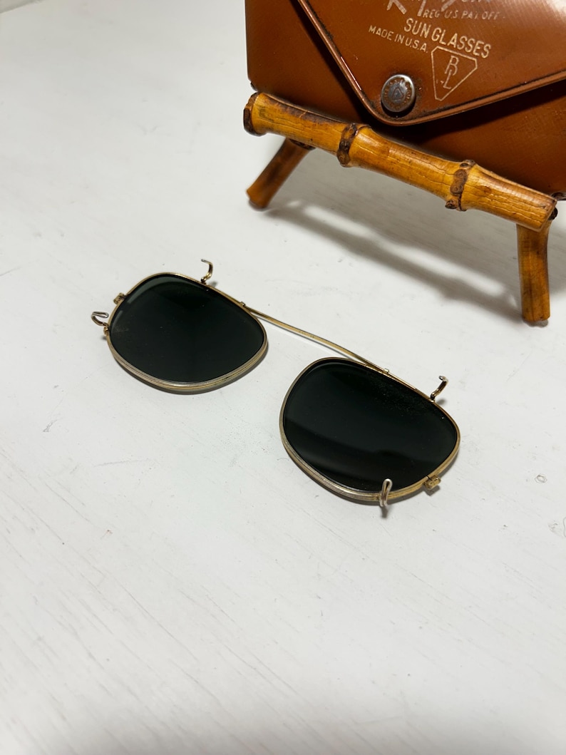 Op de afbeelding: Vintage clip-on zonnebril met donkere glazen en een goudkleurig montuur. De zonnebril ligt op een wit oppervlak, met een bruine leren etui op de achtergrond. De etui heeft een bamboe-stijl standaard.