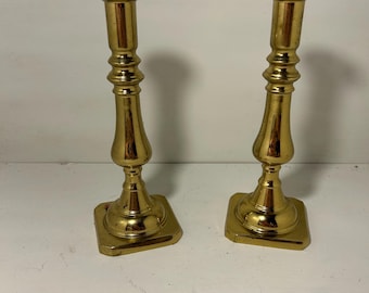 Puede incluir: Dos candeleros vintage de latón dorado. Cada candelero tiene una base cuadrada, un tallo acanalado y una parte superior en forma de copa. Los candeleros son de color dorado brillante y se exhiben sobre una superficie blanca.