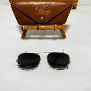 Op de afbeelding: Vintage Ray-Ban zonnebril met een bruine leren etui. De etui is gemarkeerd met "Ray-Ban Sun Glasses Made in U.S.A.". De zonnebril heeft een goudkleurig montuur en donkere glazen. De etui wordt getoond op een kleine bamboe standaard.