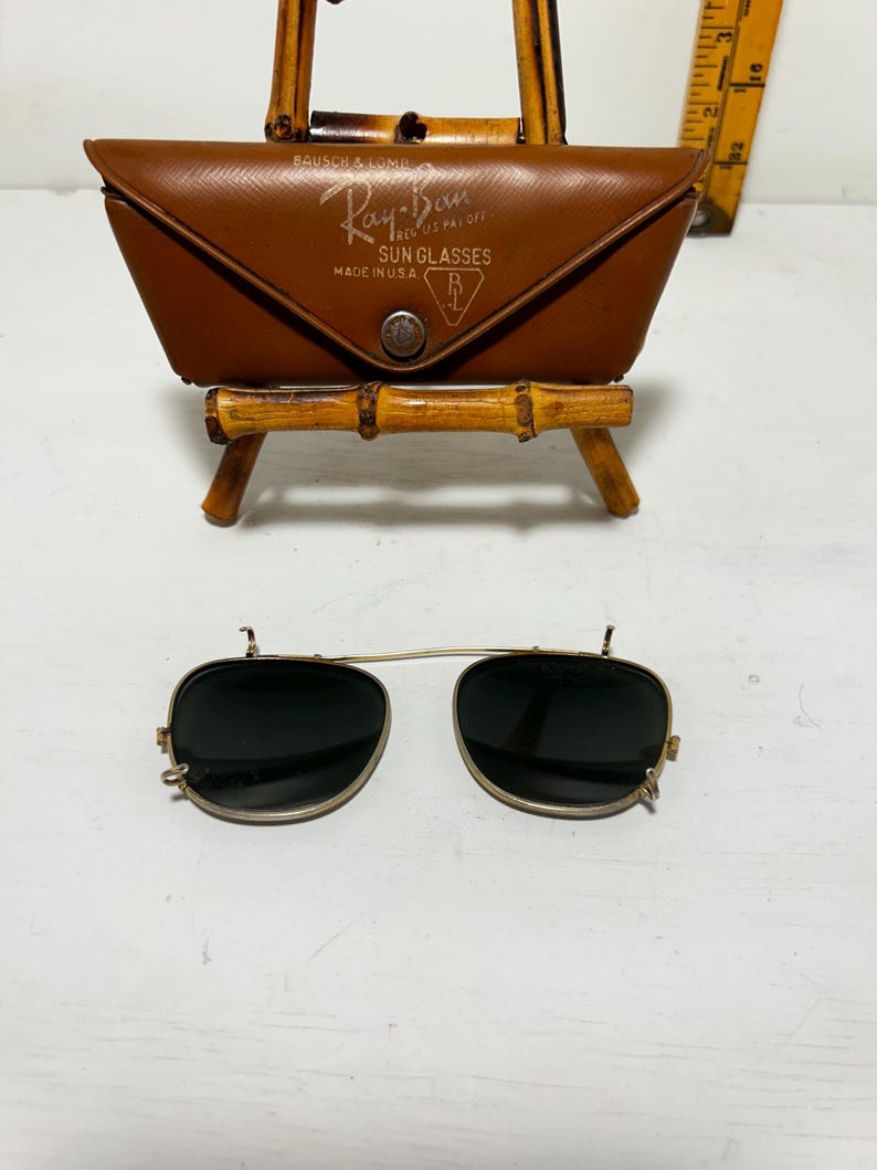 Op de afbeelding: Vintage Ray-Ban zonnebril met een bruine leren etui. De etui is gemarkeerd met "Ray-Ban Sun Glasses Made in U.S.A.". De zonnebril heeft een goudkleurig montuur en donkere glazen. De etui wordt getoond op een kleine bamboe standaard.