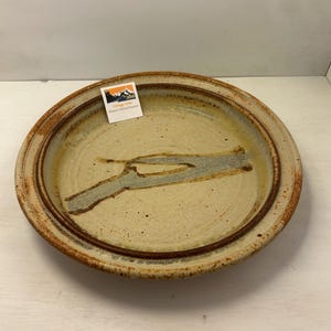 May include: A handmade ceramic plate with a beige base and a brown rim. The plate features a gray and brown abstract design in the center. A small label with a mountain logo is attached to the plate.