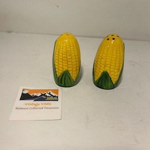 May include: A pair of ceramic salt and pepper shakers shaped like corn on the cob. The shakers are yellow with green husks and small holes on top. A small card with the text "Vintage Vista Midwest Collected Treasures" is also present.