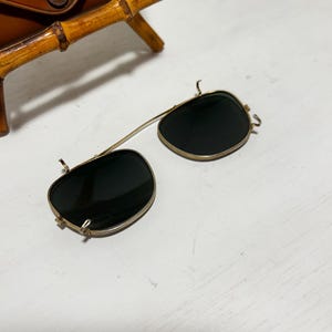 Op de afbeelding: Vintage clip-on zonnebril met donkere glazen en een goudkleurig montuur. De zonnebril ligt op een wit oppervlak, met een bruine leren etui en een arm in bamboestijl op de achtergrond. Op de etui staat "SUN GLASSES MADE IN U.S.A."