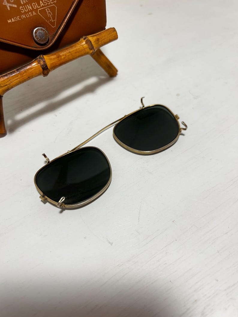 Op de afbeelding: Vintage clip-on zonnebril met donkere glazen en een goudkleurig montuur. De zonnebril ligt op een wit oppervlak, met een bruine leren etui en een arm in bamboestijl op de achtergrond. Op de etui staat "SUN GLASSES MADE IN U.S.A."