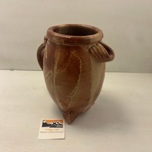 Pode incluir: Um vaso de cerâmica marrom com corpo arredondado, três pés pequenos e duas alças laterais. O vaso tem uma superfície texturizada com listras verticais de marrom mais claro. A borda é de cor marrom mais escura. Uma pequena etiqueta com texto é visível.