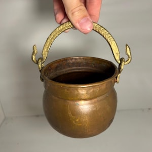 May include: A small, antique copper cauldron with a brass handle. The cauldron has a rounded body and a tarnished finish, showing signs of age. The handle is arched and attached to the cauldron with decorative brass hooks.