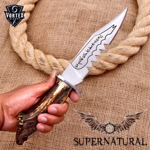 May include: A silver knife with a serrated blade and a brown antler handle. The blade has Arabic script engraved on it. The knife is held in a hand against a burlap background.