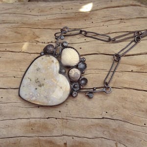 Puede incluir: Un collar artesanal con un gran colgante en forma de coraz&oacute;n con una piedra moteada de color crema. El colgante est&aacute; adornado con una concha y peque&ntilde;os adornos de metal oscuro, suspendido de una cadena de metal oscuro.