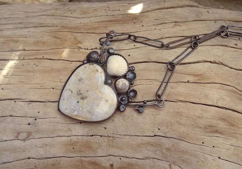 Puede incluir: Un collar artesanal con un gran colgante en forma de coraz&oacute;n con una piedra moteada de color crema. El colgante est&aacute; adornado con una concha y peque&ntilde;os adornos de metal oscuro, suspendido de una cadena de metal oscuro.