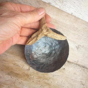 Könnte beinhalten: Eine kleine, runde Schale aus dunklem Metall mit einer gehämmerten, strukturierten Oberfläche. Die Schale verfügt über einen Griff aus einem Stück hellem, verwittertem Treibholz, das am oberen Rand des Metallbeckens befestigt ist und einen rustikalen Look erzeugt.