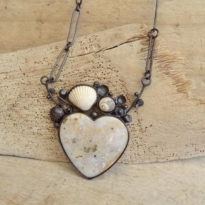 Puede incluir: Un collar artesanal con un colgante en forma de coraz&oacute;n con una superficie moteada de color blanquecino. El colgante est&aacute; enmarcado en metal oscuro y adornado con conchas marinas y peque&ntilde;os adornos redondos. El collar cuelga de una cadena oscura.