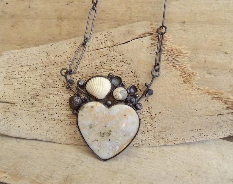 Puede incluir: Un collar artesanal con un colgante en forma de coraz&oacute;n con una superficie moteada de color blanquecino. El colgante est&aacute; enmarcado en metal oscuro y adornado con conchas marinas y peque&ntilde;os adornos redondos. El collar cuelga de una cadena oscura.