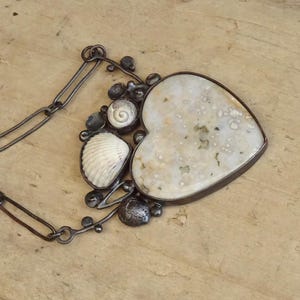 Puede incluir: Collar artesanal con un gran colgante en forma de coraz&oacute;n con una piedra moteada de color blanquecino. El colgante est&aacute; adornado con una concha, una concha en espiral y adornos de metal oscuro. El collar tiene una cadena de metal oscuro.