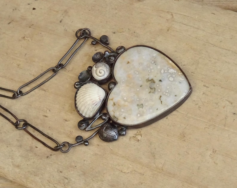 Puede incluir: Collar artesanal con un gran colgante en forma de coraz&oacute;n con una piedra moteada de color blanquecino. El colgante est&aacute; adornado con una concha, una concha en espiral y adornos de metal oscuro. El collar tiene una cadena de metal oscuro.