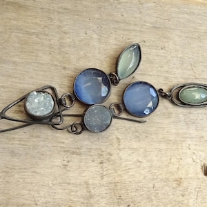 Puede incluir: Un par de pendientes colgantes con monturas de metal oscuro, piedras redondas azules facetadas, una pequeña piedra ovalada verde claro, una piedra gris circular texturizada y un marco triangular con una piedra blanca brillante.