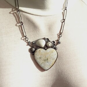 Puede incluir: Collar artesanal con un colgante en forma de coraz&oacute;n. El colgante tiene una superficie moteada de color blanquecino y est&aacute; adornado con conchas marinas y peque&ntilde;os adornos redondos. El collar tiene una cadena oscura y eslabonada.