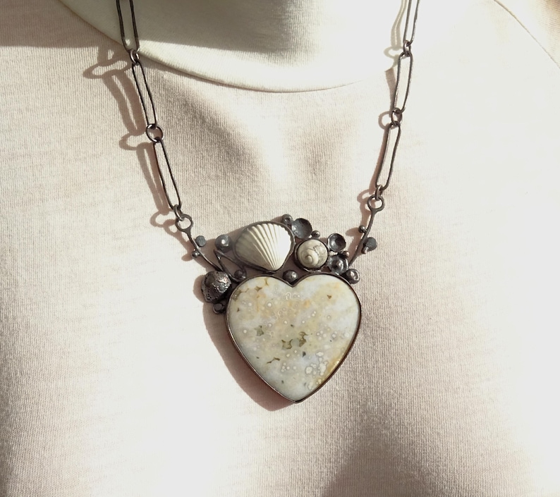 Puede incluir: Collar artesanal con un colgante en forma de coraz&oacute;n. El colgante tiene una superficie moteada de color blanquecino y est&aacute; adornado con conchas marinas y peque&ntilde;os adornos redondos. El collar tiene una cadena oscura y eslabonada.