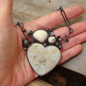 Puede incluir: Un collar artesanal con un colgante en forma de coraz&oacute;n de piedra moteada blanquecina. El colgante est&aacute; adornado con conchas marinas y detalles de metal oscuro. El collar se sostiene en una mano, mostrando el dise&ntilde;o.