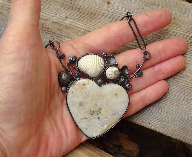 Puede incluir: Un collar artesanal con un colgante en forma de coraz&oacute;n de piedra moteada blanquecina. El colgante est&aacute; adornado con conchas marinas y detalles de metal oscuro. El collar se sostiene en una mano, mostrando el dise&ntilde;o.