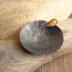 Puede incluir: Un cuenco pequeño y redondo de metal martillado con un acabado oscuro y desgastado. El cuenco presenta una pieza decorativa única en forma de lágrima, hecha de un material texturizado de color tostado claro, en el borde.