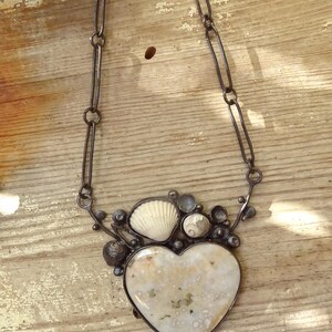 Puede incluir: Collar artesanal con un colgante en forma de coraz&oacute;n de piedra moteada color crema. El colgante est&aacute; rodeado de peque&ntilde;as conchas marinas y detalles met&aacute;licos, suspendido de una cadena.