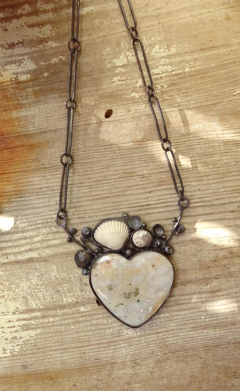 Puede incluir: Collar artesanal con un colgante en forma de coraz&oacute;n de piedra moteada color crema. El colgante est&aacute; rodeado de peque&ntilde;as conchas marinas y detalles met&aacute;licos, suspendido de una cadena.