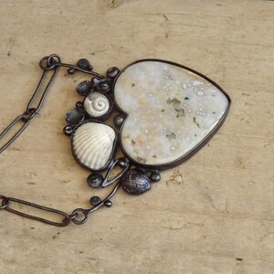 Puede incluir: Un collar artesanal con un colgante en forma de coraz&oacute;n con una piedra moteada de color blanquecino. El colgante est&aacute; enmarcado en metal oscuro y adornado con conchas marinas y cuentas oscuras. El collar tiene una cadena de metal oscuro.
