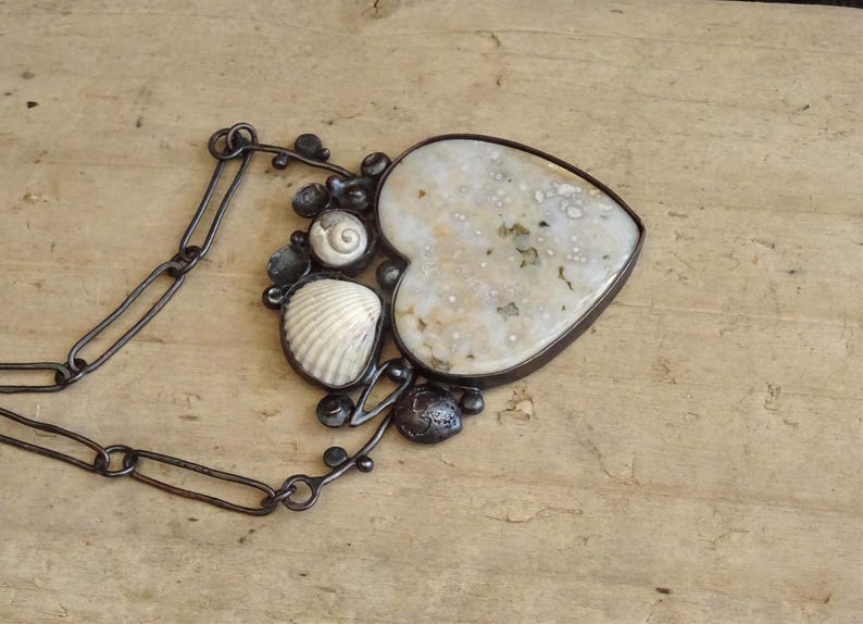 Puede incluir: Un collar artesanal con un colgante en forma de coraz&oacute;n con una piedra moteada de color blanquecino. El colgante est&aacute; enmarcado en metal oscuro y adornado con conchas marinas y cuentas oscuras. El collar tiene una cadena de metal oscuro.