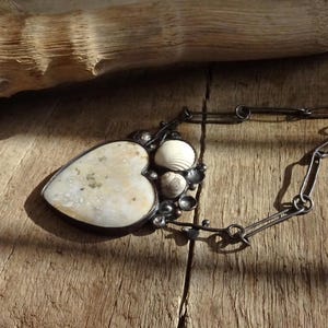Puede incluir: Un collar artesanal con un colgante en forma de coraz&oacute;n con una piedra clara y texturizada y conchas marinas. El colgante est&aacute; enmarcado en un engaste de metal oscuro y cuelga de una cadena. El collar descansa sobre una superficie de madera.