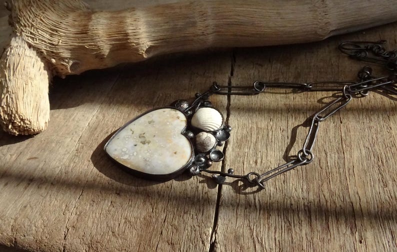 Puede incluir: Un collar artesanal con un colgante en forma de coraz&oacute;n con una piedra clara y texturizada y conchas marinas. El colgante est&aacute; enmarcado en un engaste de metal oscuro y cuelga de una cadena. El collar descansa sobre una superficie de madera.
