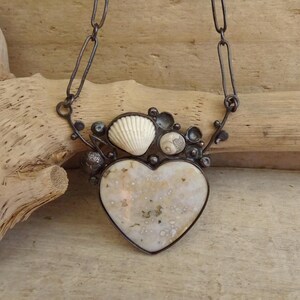 Puede incluir: Un collar artesanal con un colgante en forma de coraz&oacute;n con un patr&oacute;n moteado, adornado con conchas marinas y peque&ntilde;os elementos redondos. El colgante est&aacute; enmarcado en metal oscuro y cuelga de una cadena.