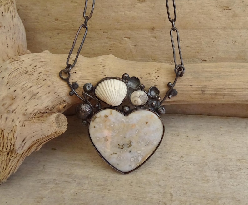 Puede incluir: Un collar artesanal con un colgante en forma de coraz&oacute;n con un patr&oacute;n moteado, adornado con conchas marinas y peque&ntilde;os elementos redondos. El colgante est&aacute; enmarcado en metal oscuro y cuelga de una cadena.