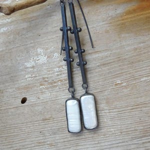 May include: A pair of long, dark metal earrings. Each earring features a rectangular, white, iridescent stone at the bottom. The earrings have a linear design with small, round accents. The background is a light-colored, weathered wooden surface.