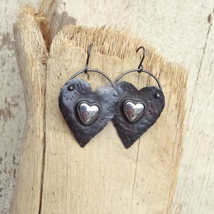 Puede incluir: Un par de pendientes en forma de corazón. Los pendientes tienen una superficie oscura y texturizada con un corazón plateado en el centro. Cuelgan de un aro circular y se exhiben sobre una superficie de madera. Joyería única.