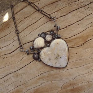 Puede incluir: Collar artesanal con un colgante en forma de coraz&oacute;n con una piedra moteada de color blanquecino. El colgante est&aacute; enmarcado en plata y adornado con conchas marinas y peque&ntilde;as cuentas oscuras. La cadena es de metal oscuro y enlazado.