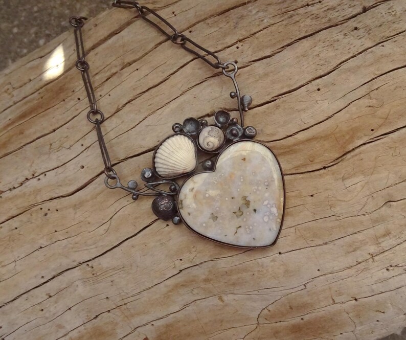 Puede incluir: Collar artesanal con un colgante en forma de coraz&oacute;n con una piedra moteada de color blanquecino. El colgante est&aacute; enmarcado en plata y adornado con conchas marinas y peque&ntilde;as cuentas oscuras. La cadena es de metal oscuro y enlazado.