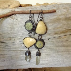 Puede incluir: Pendientes colgantes con monturas de metal oscuro, fósiles de erizo de mar en verde pálido, piedras lisas de color crema y cristales transparentes en punta. Los pendientes cuelgan de ganchos de metal oscuro, mostrando un diseño asimétrico y superpuesto con texturas naturales.