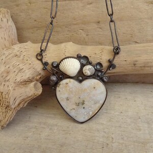 Puede incluir: Un collar artesanal con un colgante en forma de coraz&oacute;n de piedra moteada blanquecina. El colgante est&aacute; enmarcado por un marco de metal oscuro y adornado con peque&ntilde;as conchas marinas. El collar cuelga de una cadena oscura.