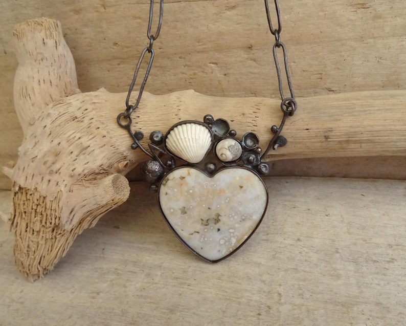 Puede incluir: Un collar artesanal con un colgante en forma de coraz&oacute;n de piedra moteada blanquecina. El colgante est&aacute; enmarcado por un marco de metal oscuro y adornado con peque&ntilde;as conchas marinas. El collar cuelga de una cadena oscura.
