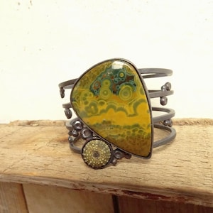 Puede incluir: Una pulsera de puño artesanal con una piedra grande de forma irregular que muestra patrones amarillos y verdes. La piedra está engastada en un marco de metal oscuro, con un pequeño acento de piedra circular. La pulsera tiene un diseño multibanda.