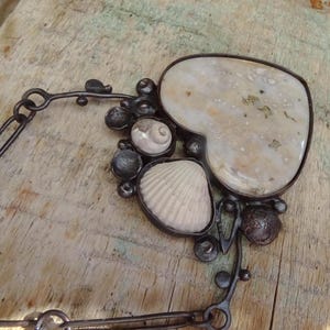 Puede incluir: Un collar artesanal con una piedra grande en forma de coraz&oacute;n y varias conchas marinas. El colgante est&aacute; enmarcado en metal oscuro e incluye una cadena. El collar est&aacute; sobre un fondo de madera.