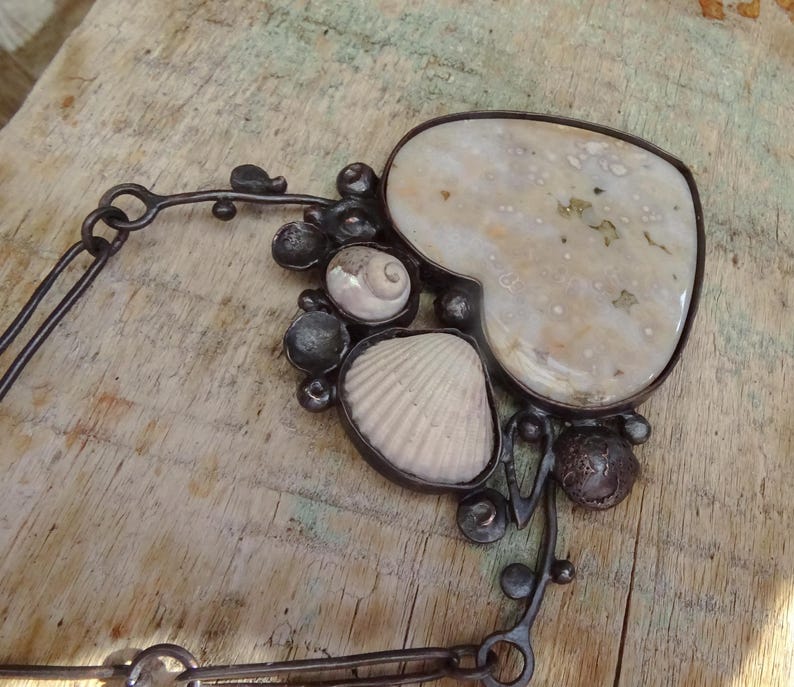 Puede incluir: Un collar artesanal con una piedra grande en forma de coraz&oacute;n y varias conchas marinas. El colgante est&aacute; enmarcado en metal oscuro e incluye una cadena. El collar est&aacute; sobre un fondo de madera.