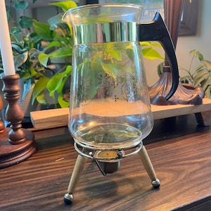May include: A vintage glass coffee carafe with a black handle and gold-colored trim. The carafe sits on a metal and wood stand with three legs. The glass has a decorative pattern.