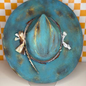 May include: A turquoise fedora hat with a distressed finish. The hat has a decorative band with feathers and fabric accents. The brim and crown have a mottled, aged appearance with gold and brown highlights, giving it a vintage aesthetic.