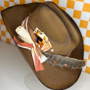 May include: A brown cowboy hat with a decorative band featuring fabric, feathers, and playing cards. The hat has a wide brim and a rounded crown. The decorative elements include a gray feather and a playing card.